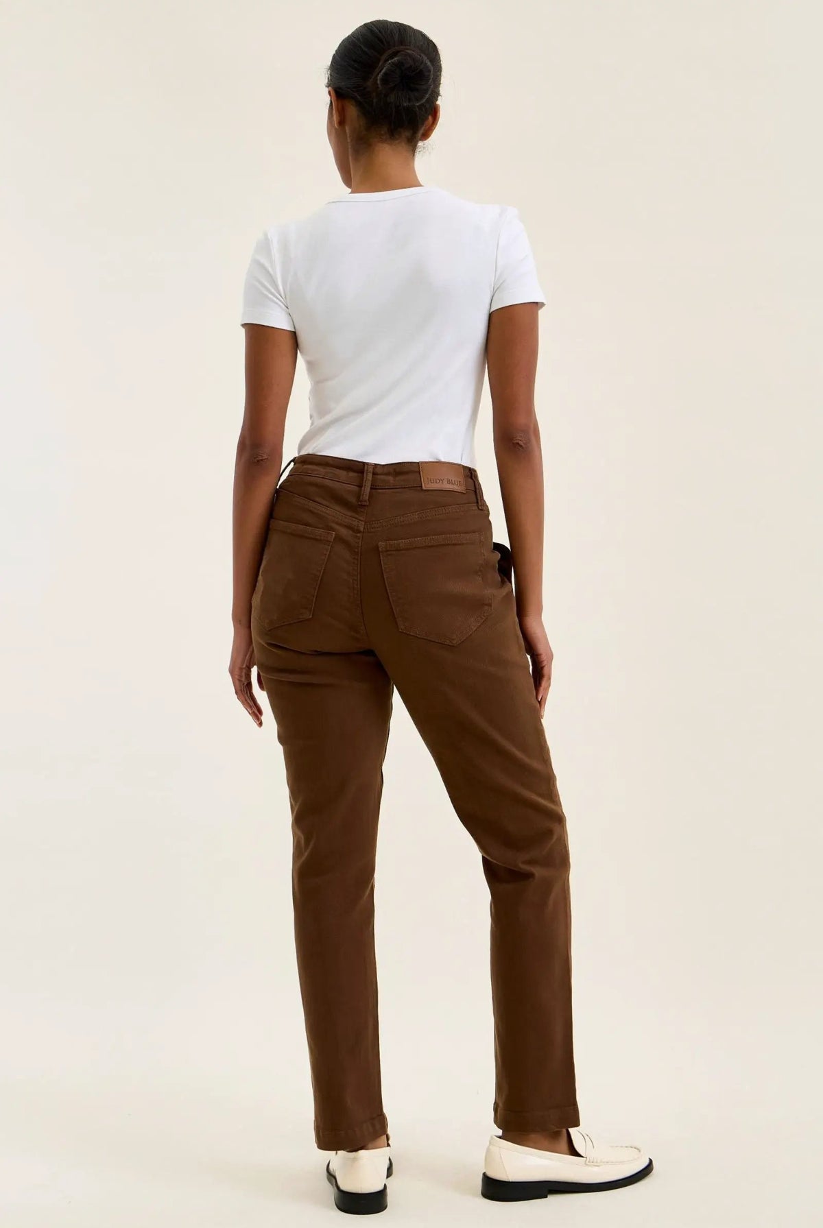 Back view of mahogany high-waist stretch jeans with a slim straight leg, fitted top block, and double-rolled cuff paired with white loafers.