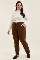 Plus model wearing mahogany high-waist stretch jeans with utility pockets and slim-straight legs, paired with a cream top.