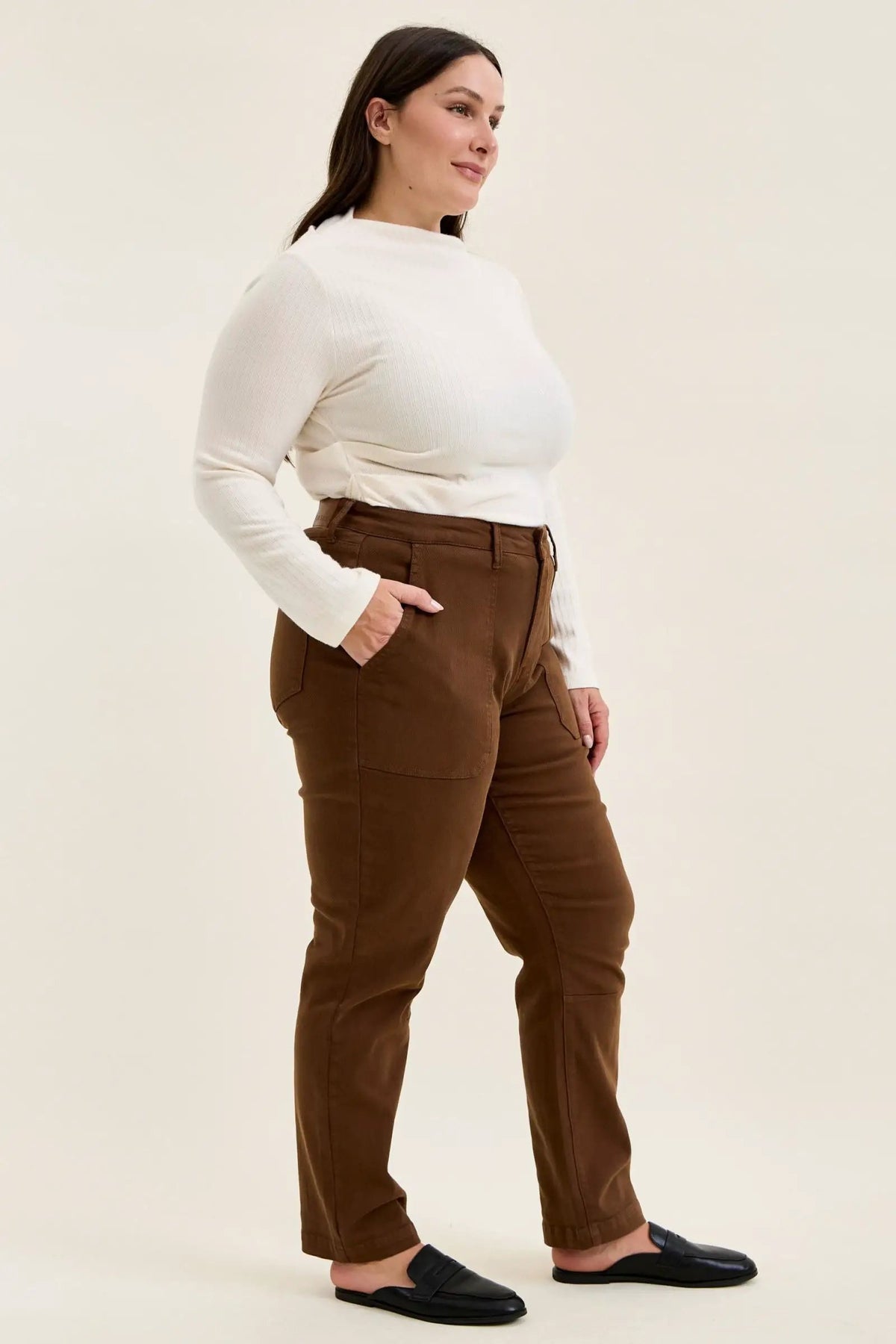 Plus-size model wearing mahogany high-waist stretch jeans with utility pockets and slim-straight legs, styled with a white top and black loafers.