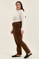 Plus model wearing mahogany high-waist stretch jeans with utility pockets and double-rolled cuffs, paired with a white top and black mules.