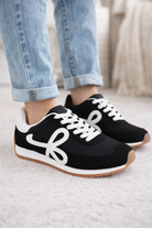 Black retro low top sneakers with white side logo, white laces, and gum sole worn with cuffed blue jeans.